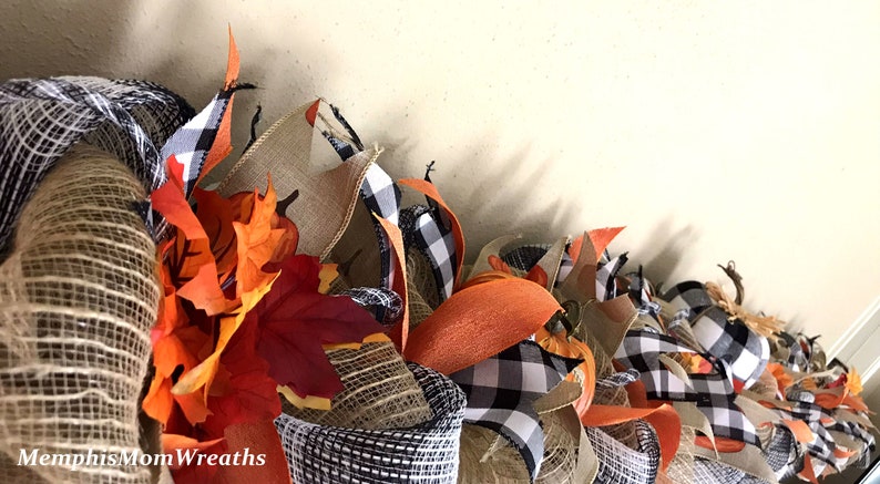 Puede incluir: Una guirnalda decorativa con malla de arpillera de color natural, cinta de cuadros negros y blancos y hojas de arce artificiales de oto&ntilde;o en tonos naranja, rojo y amarillo. El texto MemphisMomWreaths est&aacute; impreso en blanco en la esquina.