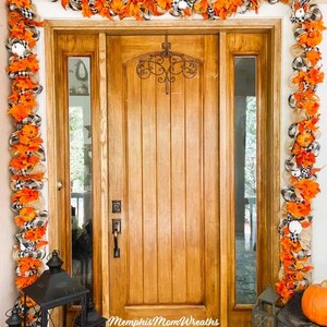 Puede incluir: Una puerta de entrada de madera enmarcada por una guirnalda decorativa con hojas artificiales naranjas, malla de arpillera y cintas de cuadros negros y blancos. Un colgador de metal negro est&aacute; fijado en la puerta. El texto Memphis Mom Wreaths es visible.