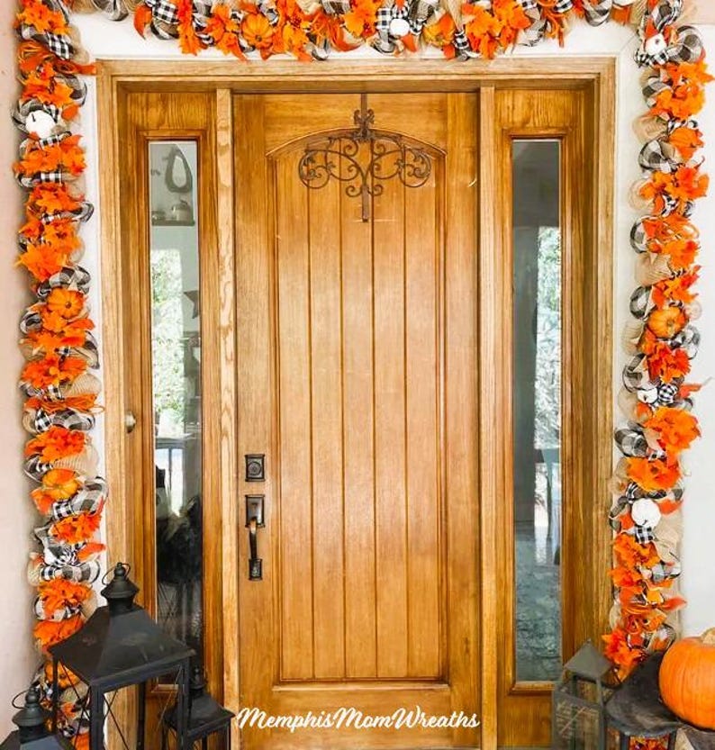 Puede incluir: Una puerta de entrada de madera enmarcada por una guirnalda decorativa con hojas artificiales naranjas, malla de arpillera y cintas de cuadros negros y blancos. Un colgador de metal negro est&aacute; fijado en la puerta. El texto Memphis Mom Wreaths es visible.