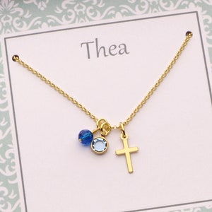 Puede incluir: Un collar de cadena dorada con un pequeño dije de cruz y dos cuentas de cristal azul. El collar se muestra en una tarjeta blanca con el nombre "Thea" impreso en ella.