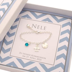 May include: A silver bracelet with a turquoise crystal, angel charm, and a personalised disc that says "Nele". The bracelet is in a white box with a blue and white chevron pattern. The box also includes the text "Zu deiner Kommunion am 27. April 2025".