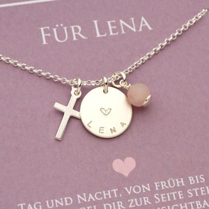 Könnte beinhalten: Silberkette mit einem Kreuzanhänger, einem runden Anhänger mit dem Namen "Lena" und einem Herz sowie einer rosafarbenen Edelsteinperle. Die Kette liegt auf einem rosa Untergrund mit dem Text "Für Lena" und einem kleinen Herz.