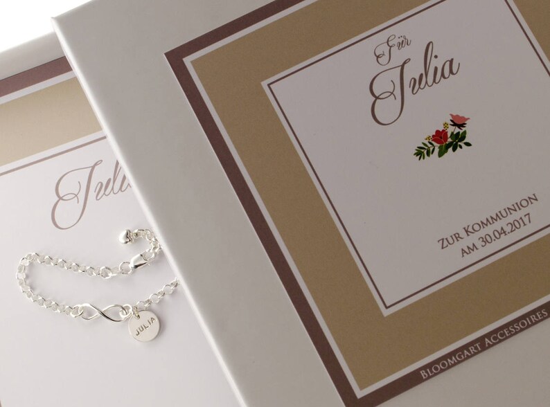 May include: A white gift box with a brown border and the text "F&uuml;r Julia Zur Kommunion am 30.04.2017" and the text "BLOOMGART ACCESSOIRES". A silver bracelet with a charm that says "Julia" is on the box.