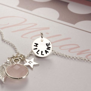 Puede incluir: Un collar de plata con un colgante redondo que dice "MILANE" en letras negras. El collar tambi&eacute;n tiene un dije de cristal rosa, un dije de estrella plateada con la letra "M" y un dije de ala de &aacute;ngel plateada.