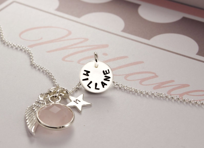 Puede incluir: Un collar de plata con un colgante redondo que dice "MILANE" en letras negras. El collar tambi&eacute;n tiene un dije de cristal rosa, un dije de estrella plateada con la letra "M" y un dije de ala de &aacute;ngel plateada.