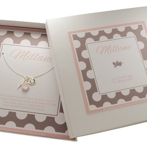 Puede incluir: Una caja de regalo blanca con un patr&oacute;n de lunares rosa y marr&oacute;n. La caja contiene un collar de plata con una piedra rosa, un dije de pluma y un peque&ntilde;o disco de plata. La tapa de la caja tiene el nombre "Millane" y el texto "Gemelas nacidas el 21 de marzo de 2014".