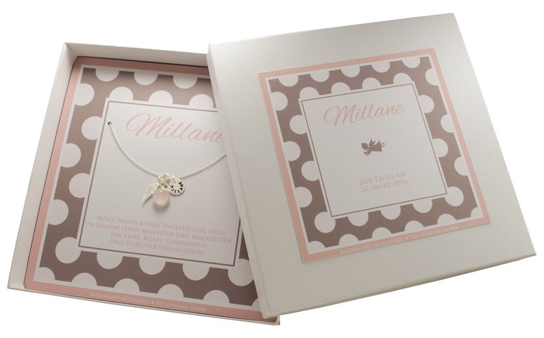 Puede incluir: Una caja de regalo blanca con un patr&oacute;n de lunares rosa y marr&oacute;n. La caja contiene un collar de plata con una piedra rosa, un dije de pluma y un peque&ntilde;o disco de plata. La tapa de la caja tiene el nombre "Millane" y el texto "Gemelas nacidas el 21 de marzo de 2014".