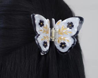 May include: A white butterfly hair clip with black floral details and gold accents. The clip is made of plastic and has a spring-loaded mechanism.