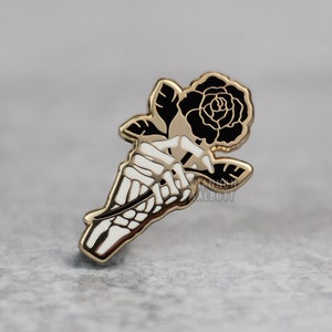 May include: A gold-colored enamel pin featuring a black rose held by a white skeleton hand. The pin has a gold outline and is set against a gray background. The design is detailed and gothic-inspired.
