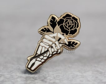 May include: A gold-colored enamel pin featuring a black rose held by a white skeleton hand. The pin has a gold outline and is set against a gray background. The design is detailed and gothic-inspired.