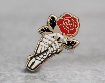 May include: A gold-colored enamel pin featuring a skeleton hand holding a red rose. The rose has black and gold-colored leaves. The pin has a gold border and is set against a gray background.