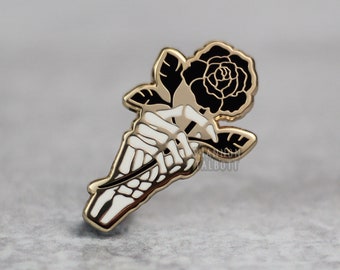 May include: A gold-colored enamel pin featuring a black rose held by a white skeleton hand. The pin has a detailed design with a gold outline and is set against a gray background. The rose has a detailed petal design.
