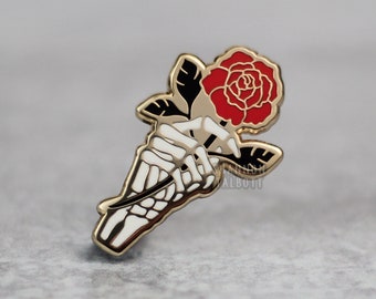 May include: A gold-colored enamel pin featuring a skeletal hand holding a red rose. The rose has green leaves and a black outline. The pin has a gold border and is set against a gray background.