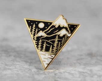 May include: Gold-tone enamel pin featuring a black and white mountain scene with a crescent moon and stars. The pin is shaped like a triangle.