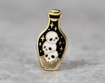 Puede incluir: Pin de esmalte dorado con una botella negra llena de tres calaveras blancas y purpurina dorada. La botella tiene un tapón dorado y está sobre un fondo negro.