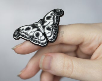 以下が含まれることがあります： 蝶のデザインが施された黒と白のエナメルピン。ピンは指につけられています。