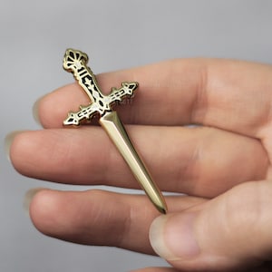 May include: A gold-colored dagger pin with a cross-shaped hilt and a pointed blade. The hilt features intricate black and white detailing. The pin is held in a person's hand.