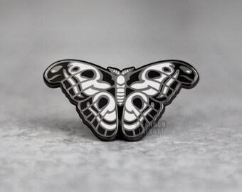 以下が含まれることがあります： 黒と白のエナメルピンで、蝶のデザインが細かく描かれています。蝶は白い体と、複雑な模様の黒い羽を持っています。