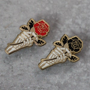 May include: Two enamel pins featuring a skeleton hand holding a rose. One rose is red, the other is black. The pins have a gold outline and white bone details. The artist's name, SHANNON TALBOTT, is printed on the pin.