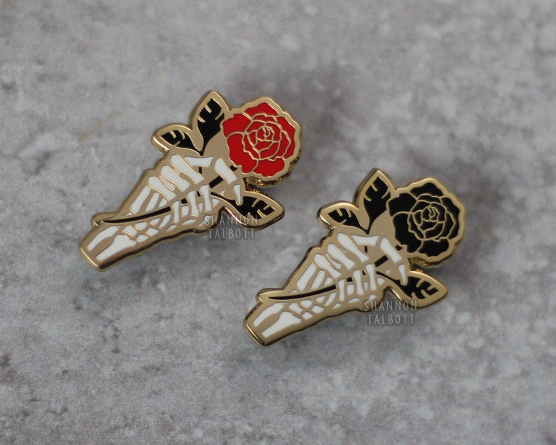 May include: Two enamel pins featuring a skeleton hand holding a rose. One rose is red, the other is black. The pins have a gold outline and white bone details. The artist's name, SHANNON TALBOTT, is printed on the pin.