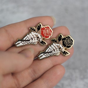 May include: Two enamel pins featuring a skeleton hand holding a rose. One rose is red, the other is black. Both pins have gold-colored borders and black and white details. The pins are displayed against a neutral background.
