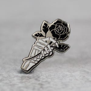 K&ouml;nnte beinhalten: Ein silberfarbener Ansteckpin mit einer Skeletthand, die eine schwarze Rose h&auml;lt. Die Rose hat detaillierte Bl&uuml;tenbl&auml;tter, und der Pin hat eine polierte Oberfl&auml;che. Der Name des K&uuml;nstlers, SHANNON TALBOTT, ist auf dem Pin aufgedruckt.
