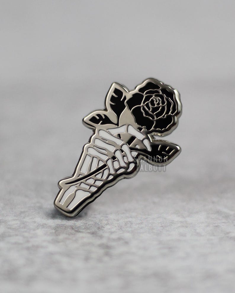 K&ouml;nnte beinhalten: Ein silberfarbener Ansteckpin mit einer Skeletthand, die eine schwarze Rose h&auml;lt. Die Rose hat detaillierte Bl&uuml;tenbl&auml;tter, und der Pin hat eine polierte Oberfl&auml;che. Der Name des K&uuml;nstlers, SHANNON TALBOTT, ist auf dem Pin aufgedruckt.
