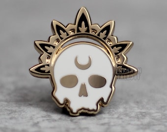 May include: A gold-coloured metal pin featuring a white skull with a crescent moon and floral accents. The skull is centered within a gold sunburst design. The pin is likely an enamel or hard-wearing metal.