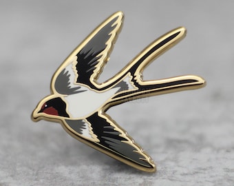 Puede incluir: Un pin de metal dorado con forma de golondrina en vuelo. El ave tiene plumas negras, blancas y grises, con una mancha roja en la cabeza. El pin está detallado con un contorno dorado.