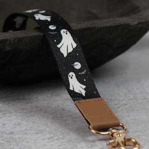 May include: Black and white ghost patterned lanyard with a brown leather strap and a gold metal clasp.