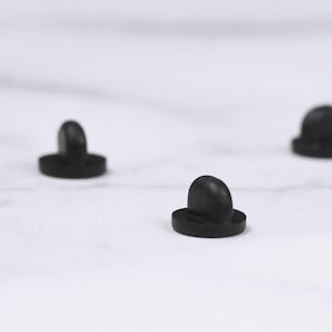 May include: Close-up of three small, black, mushroom-shaped objects. Each object has a rounded top and a flat, circular base. The objects are evenly spaced against a white background.
