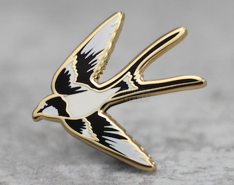 Puede incluir: Pin de esmalte dorado con una golondrina negra y blanca en vuelo. La golondrina tiene el vientre blanco y las alas negras con detalles blancos.