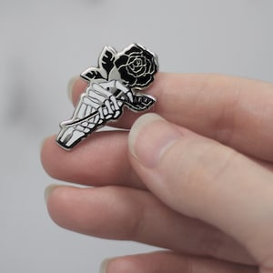 K&ouml;nnte beinhalten: Ein silberfarbener Emaille-Pin mit einer Skeletthand, die eine schwarze Rose h&auml;lt. Die Rose hat detaillierte Bl&uuml;tenbl&auml;tter und gr&uuml;ne Bl&auml;tter. Ein Accessoire im Gothic-Stil.