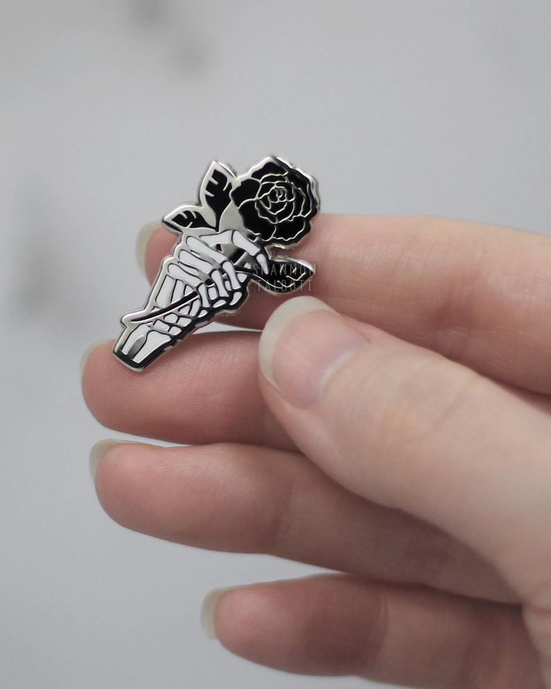 K&ouml;nnte beinhalten: Ein silberfarbener Emaille-Pin mit einer Skeletthand, die eine schwarze Rose h&auml;lt. Die Rose hat detaillierte Bl&uuml;tenbl&auml;tter und gr&uuml;ne Bl&auml;tter. Ein Accessoire im Gothic-Stil.
