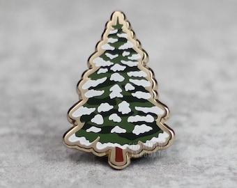 May include: A gold-colored metal pin in the shape of a Christmas tree. The tree is green with white snow accents and a brown trunk. The pin has a gold border and is set against a gray background.
