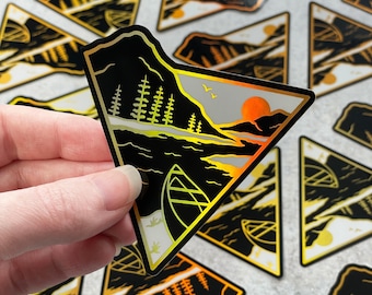 May include: A holographic sticker featuring a black and gold illustration of a mountain lake scene with a canoe, trees, and a sunset. The sticker is shaped like a triangle.