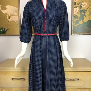 May include: A navy blue and white gingham dress with a red trim around the neckline and buttons. The dress has a fitted bodice and a full skirt. It has a belt at the waist.