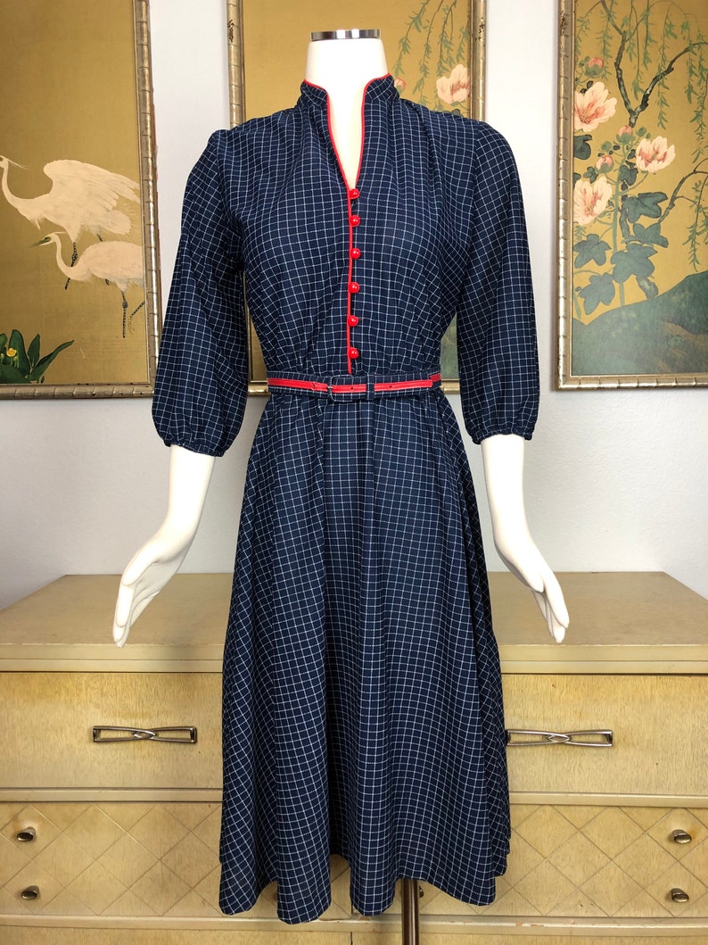 May include: A navy blue and white gingham dress with a red trim around the neckline and buttons. The dress has a fitted bodice and a full skirt. It has a belt at the waist.