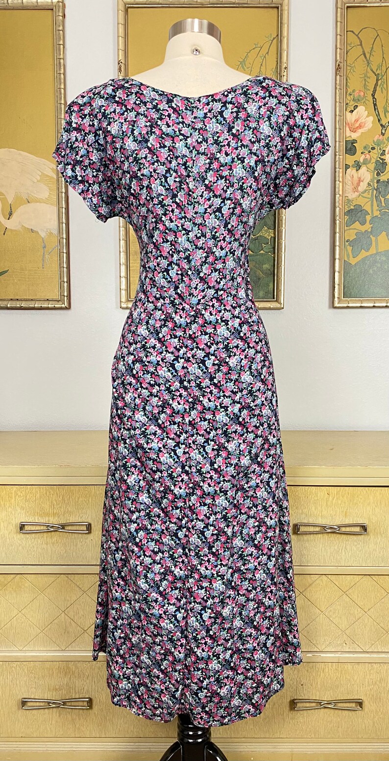 Może przedstawiać: Sukienka w stylu vintage z kwiatowym nadrukiem na manekinie. Sukienka ma ciemne tło z wzorem małych r&oacute;żowych, niebieskich i białych kwiat&oacute;w. Ma kr&oacute;tkie rękawy i sp&oacute;dnicę o długości midi, prezentując klasyczny, kobiecy styl.