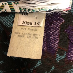 May include: A white tag with black text that reads "Size 14", "100% Rayon", "Dry Clean Only", and "Made in USA". The tag is attached to a black fabric with a colorful pattern.