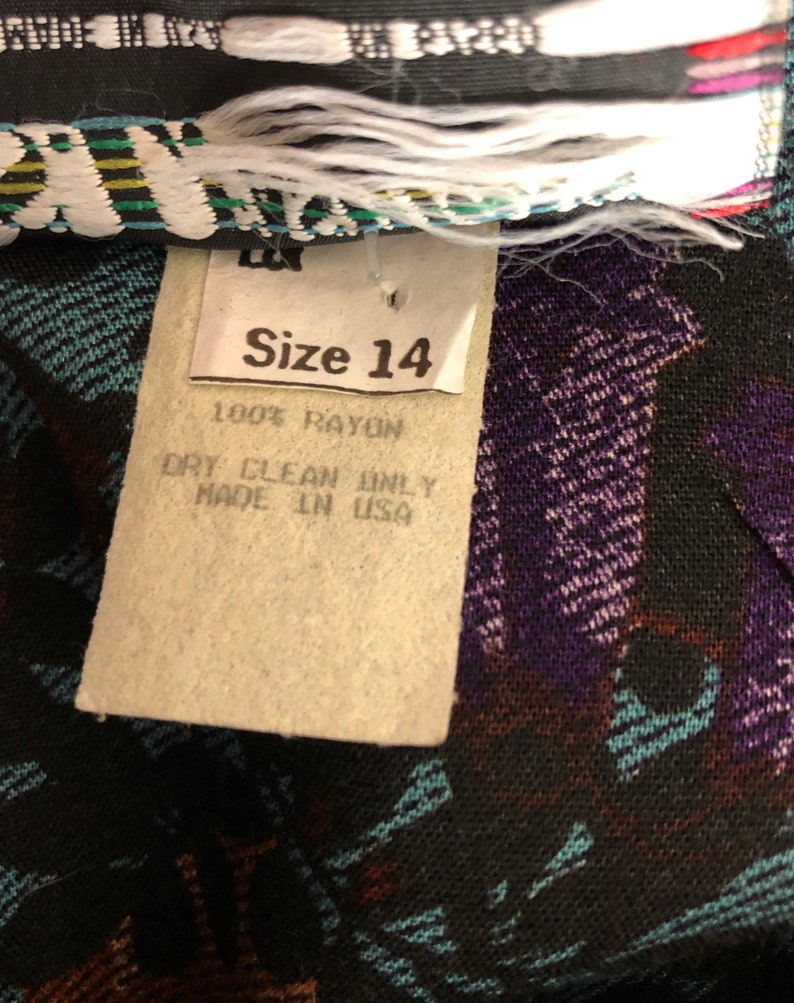May include: A white tag with black text that reads "Size 14", "100% Rayon", "Dry Clean Only", and "Made in USA". The tag is attached to a black fabric with a colorful pattern.