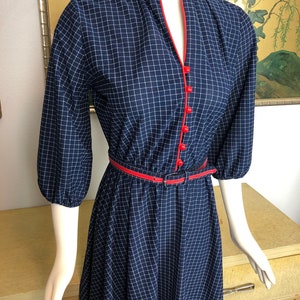 May include: A navy blue and white gingham dress with a red trim around the neckline and a black belt. The dress has a v-neck and 3/4 sleeves.