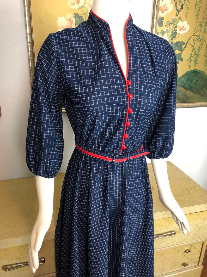 May include: A navy blue and white gingham dress with a red trim around the neckline and a black belt. The dress has a v-neck and 3/4 sleeves.