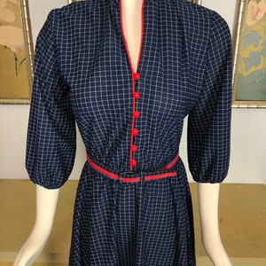 May include: A navy blue dress with a white gingham pattern. The dress has a red trim around the neckline and a red belt. The dress has a v-neck and 3/4 sleeves.