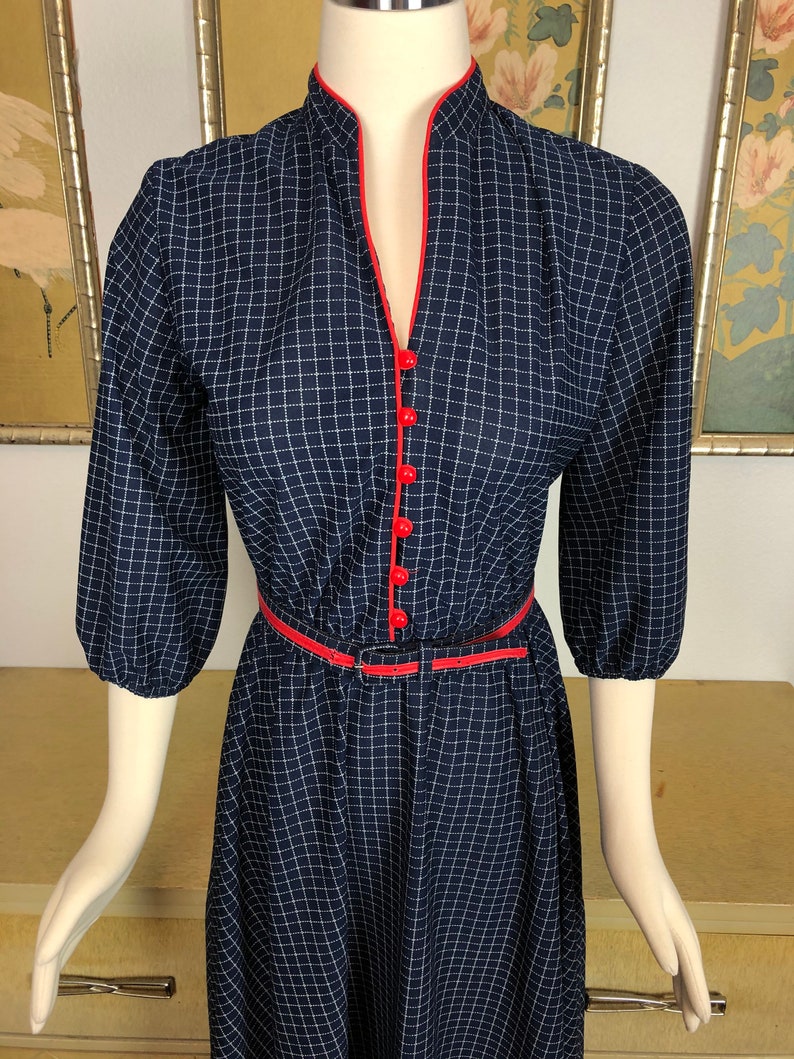 May include: A navy blue dress with a white gingham pattern. The dress has a red trim around the neckline and a red belt. The dress has a v-neck and 3/4 sleeves.