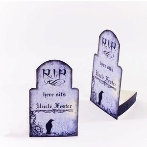 Puede incluir: Dos tarjetas de lugar de Halloween en forma de l&aacute;pida, de color morado y negro, con el texto "R.I.P. here sits Uncle Fester" y una ilustraci&oacute;n de cuervo negro.