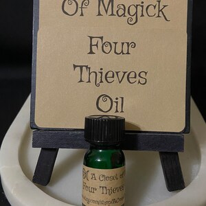 May include: A small green glass bottle with a black cap, labeled "A Closet of Four Thieves Oil" on a white surface. The bottle is in front of a brown sign with the text "A Closet of Magick Four Thieves Oil" and a triple moon symbol.