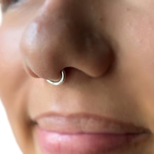 Puede incluir: Primer plano de un aro de nariz plateado. El aro tiene un diseño simple y circular, y se usa en el piercing del tabique nasal. La piel alrededor de la nariz es visible, con un tono suave y natural. La atención se centra en la joya.
