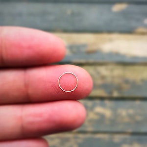 Puede incluir: Un pequeño pendiente de aro de color dorado. El pendiente tiene una forma circular simple y se sostiene entre dos dedos. El fondo es una superficie de madera borrosa.