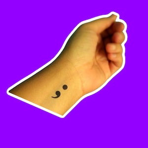 May include: A black semicolon tattoo on the wrist. The tattoo is a small, simple design, and the skin has a light brown tone. The background is a vibrant purple color, with a white border around the arm.